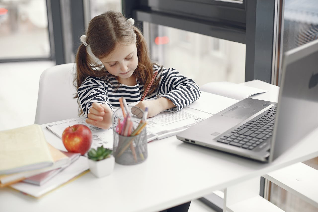 A young girl studies at home using a laptop, engaged with her homework and school supplies.